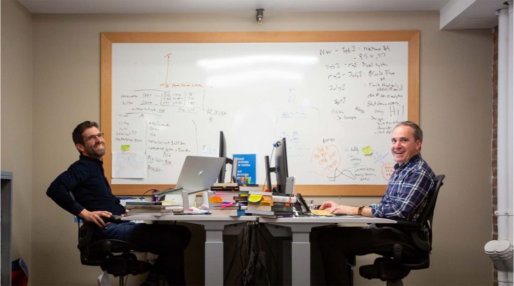 Gordon Ross and Darren Gibbons sitting at a shared desk laughing together in the office.