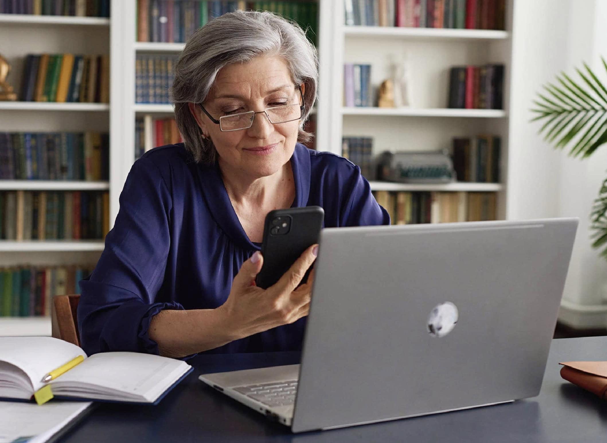An older woman sitting at a desk with a laptop and legal books behind her, making a call on her smartphone