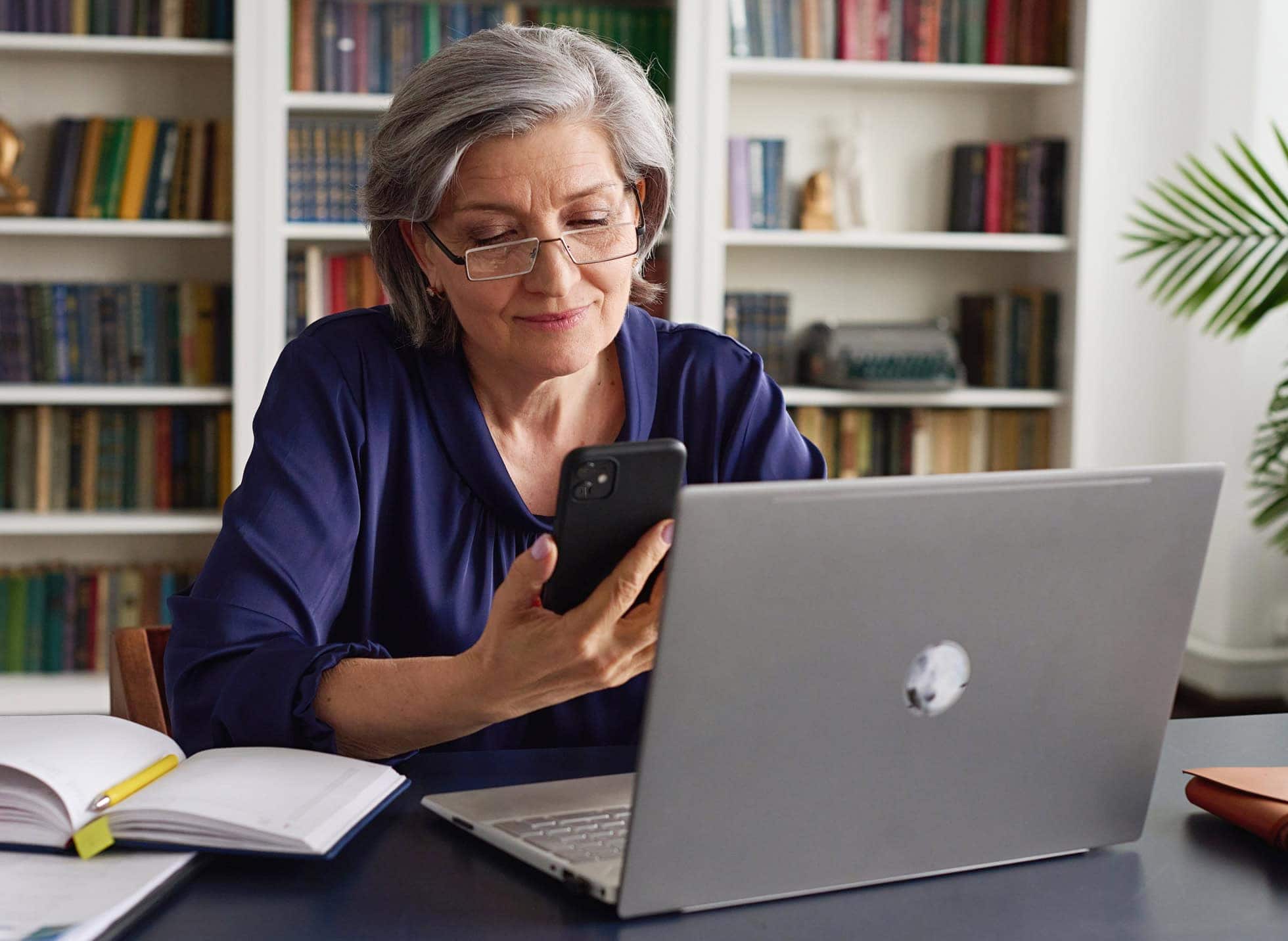 An older woman sitting at a desk with a laptop and legal books behind her, making a call on her smartphone