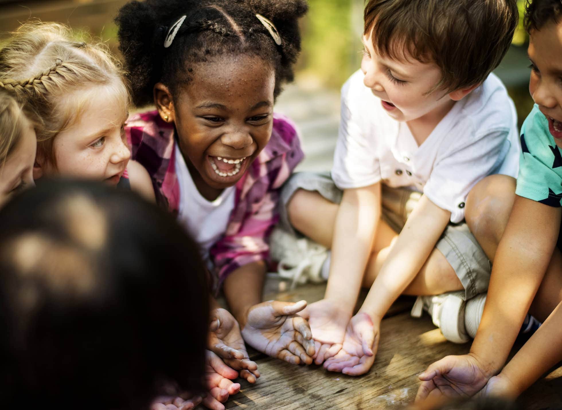 A diverse group of children laughing together and holding out their hands while sitting in a circle outdoors